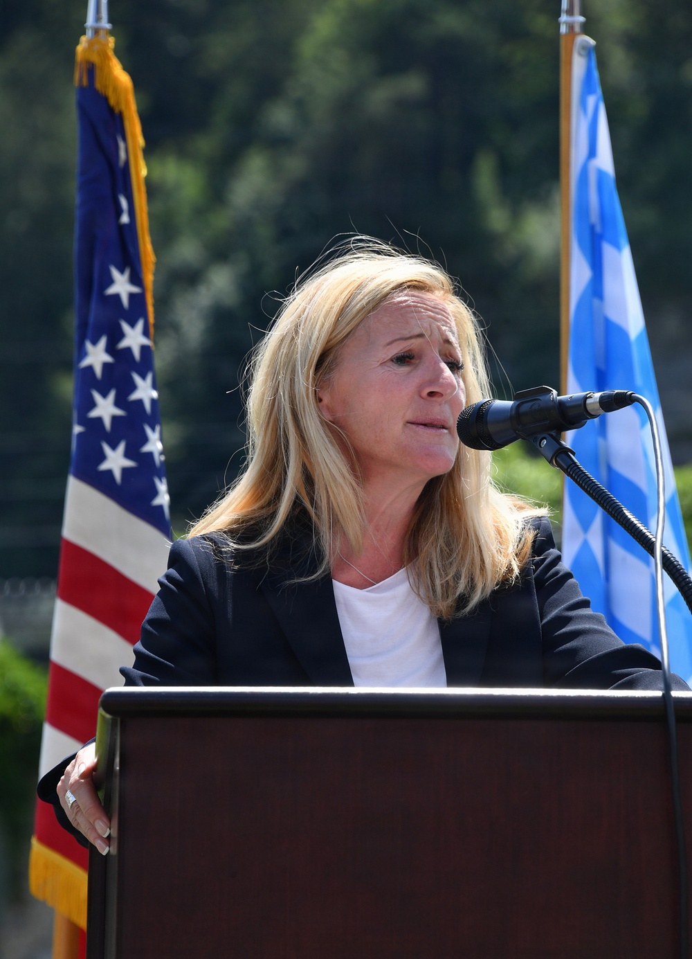 A woman with blonde hair is speaking at a wooden podium with a microphone at an outdoor event. She is wearing a dark blazer over a white shirt and appears to be delivering a speech. Two flags are visible behind her — a United States flag on the left and what appears to be a Greek flag on the right.