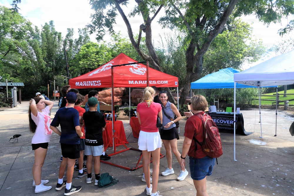 An outdoor community event featuring a U.S. Marines recruitment tent (red canopy with 'MARINES' and 'MARINES.COM' branding) alongside a 'Hillsborough Recovery Coalition' booth under a blue and white canopy. A group of approximately 8-10 people, including youth and adults, are gathered in what appears to be a park or zoo setting. The event takes place on a paved plaza area surrounded by trees and natural rock formations.