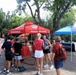 An outdoor community event featuring a U.S. Marines recruitment tent (red canopy with 'MARINES' and 'MARINES.COM' branding) alongside a 'Hillsborough Recovery Coalition' booth under a blue and white canopy. A group of approximately 8-10 people, including youth and adults, are gathered in what appears to be a park or zoo setting. The event takes place on a paved plaza area surrounded by trees and natural rock formations.