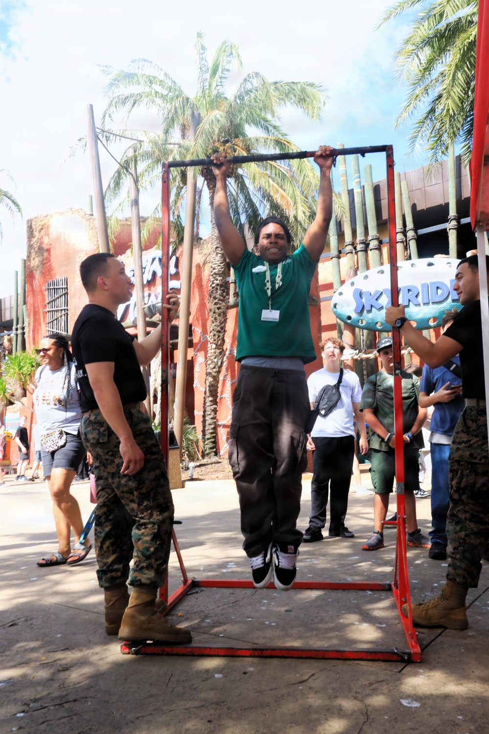 A civilian man in a green t-shirt and black cargo pants is performing pull-ups on a portable red metal pull-up bar frame at what appears to be a theme park or outdoor event. Military personnel in USMC MARPAT camouflage uniforms are present on both sides of the frame, observing and supervising the activity. A crowd of onlookers is gathered in the background near a 'Sky Ride' attraction sign surrounded by palm trees.