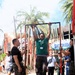 A civilian man in a green t-shirt and black cargo pants is performing pull-ups on a portable red metal pull-up bar frame at what appears to be a theme park or outdoor event. Military personnel in USMC MARPAT camouflage uniforms are present on both sides of the frame, observing and supervising the activity. A crowd of onlookers is gathered in the background near a 'Sky Ride' attraction sign surrounded by palm trees.