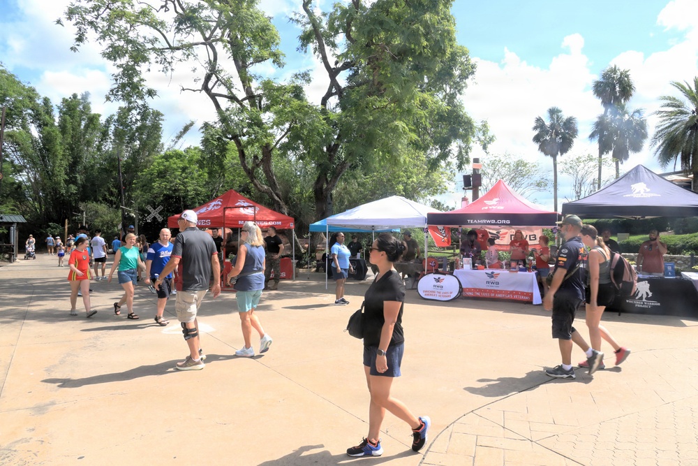 An outdoor event or fair is taking place in what appears to be a park or theme park setting, featuring multiple vendor/organization tents. Visible organizations include the U.S. Marines (marines.com), Team RWB (teamrwb.org), and what appears to be the Wounded Warrior Project, suggesting a military/veterans appreciation event. Numerous visitors are walking through the area under partly cloudy skies surrounded by lush trees.