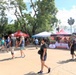 An outdoor event or fair is taking place in what appears to be a park or theme park setting, featuring multiple vendor/organization tents. Visible organizations include the U.S. Marines (marines.com), Team RWB (teamrwb.org), and what appears to be the Wounded Warrior Project, suggesting a military/veterans appreciation event. Numerous visitors are walking through the area under partly cloudy skies surrounded by lush trees.