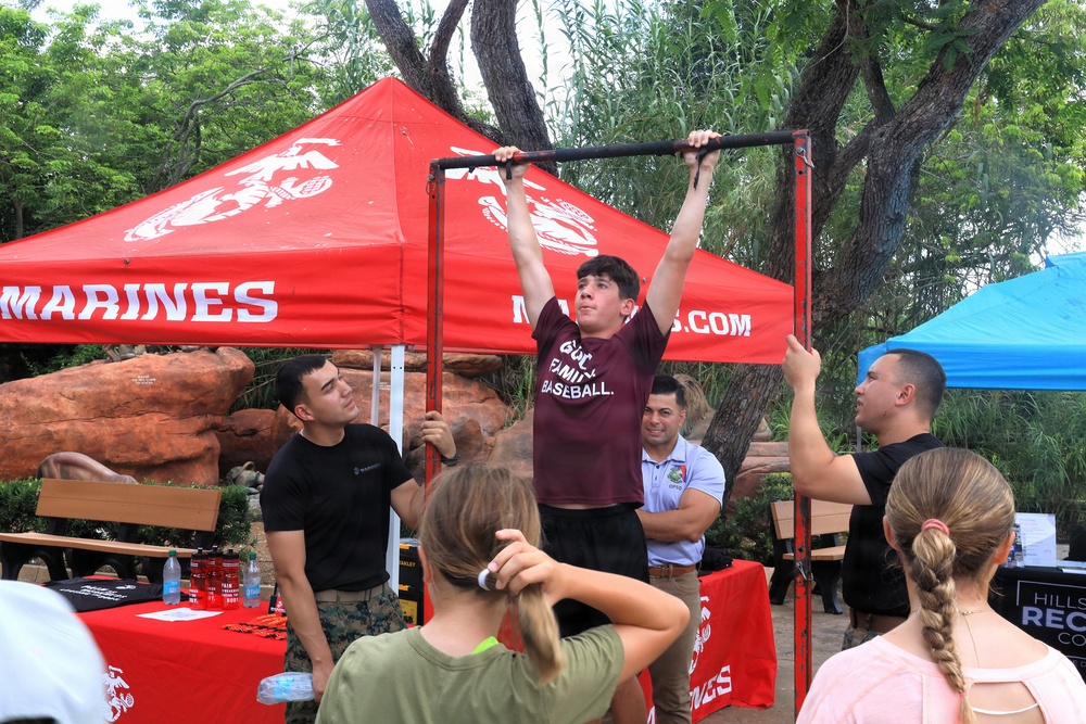 A young person in a maroon 'God. Family. Baseball.' t-shirt is performing pull-ups on a portable bar at a U.S. Marines recruiting booth. Two Marines in black uniforms are observing and assisting, while a small crowd watches. The event appears to be an outdoor recruitment or community fair set up under red Marines-branded canopy tents.