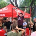 A young person in a maroon 'God. Family. Baseball.' t-shirt is performing pull-ups on a portable bar at a U.S. Marines recruiting booth. Two Marines in black uniforms are observing and assisting, while a small crowd watches. The event appears to be an outdoor recruitment or community fair set up under red Marines-branded canopy tents.