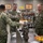 Three U.S. Navy personnel in NWU Type III camouflage uniforms are gathered around a workbench in what appears to be a technical training facility or workshop. A sailor facing away from the camera appears to be briefing or demonstrating equipment to two senior officers, one identified by nametape as 'SHELDON' and another as 'FOSTER', both wearing U.S. Navy insignia. The setting suggests a maintenance, engineering, or damage control training environment.