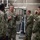 A group of U.S. Navy personnel in Navy Working Uniform (NWU) Type III camouflage are gathered inside what appears to be a mechanical or utility room. One individual, whose name tape reads 'SHELDON' and bears U.S. Navy insignia, appears to be briefing or explaining something to the group. The setting suggests a facility inspection or operational tour.