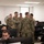A group of military personnel in U.S. Navy Working Uniform (NWU Type III) camouflage are gathered in what appears to be a computer lab or operations center. Several uniformed service members are engaged in conversation in the center of the room while others, likely trainees or operators, are seated at computer workstations. The setting suggests a training facility, briefing room, or cyber operations environment.