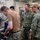 Four U.S. Navy personnel are gathered in what appears to be a machinist or fabrication shop, observing work being performed on a metal lathe or similar machine tool. The individuals are wearing safety goggles and a mix of Navy Working Uniforms (NWU) and a tan 'Machinist Mate' t-shirt, suggesting a training or inspection scenario. The setting is an industrial workshop with multiple machines visible in the background.