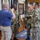 A group of U.S. Navy personnel in NWU Type III (woodland camouflage) uniforms are gathered in what appears to be a military building lobby or reception area. A civilian in a blue polo shirt and khaki pants is engaged in conversation with several uniformed sailors. The setting includes a 'WELCOME' mosaic floor mat, an American flag, and official Navy insignia/seals on the walls.