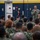 A senior U.S. Navy officer or senior enlisted leader is addressing a large group of military personnel in what appears to be a hangar bay or indoor military facility. The speaker is holding a microphone and speaking to a seated audience of sailors and possibly Marines in various camouflage uniforms. The setting suggests an all-hands meeting, command briefing, or official address.