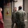 A group of U.S. Navy personnel in digital camouflage uniforms are engaged in a conversation with a civilian or technician wearing a red jumpsuit inside what appears to be a military training or maintenance facility. The setting includes concrete block walls, red fire suppression piping, a fire extinguisher mounted in a yellow-and-black hazard-marked cabinet, and a sign reading 'INSTRUCTOR USE ONLY.' The interaction suggests a briefing, inspection, or training walkthrough.
