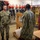 Three military personnel are gathered in what appears to be a fire station equipment room on a U.S. Navy installation. Two individuals are wearing U.S. Navy NWU Type III (digital camouflage) uniforms, while a third person is wearing red coveralls. The room is equipped with firefighting gear including red helmets, SCBA masks, and turnout/bunker coats hanging on the walls.
