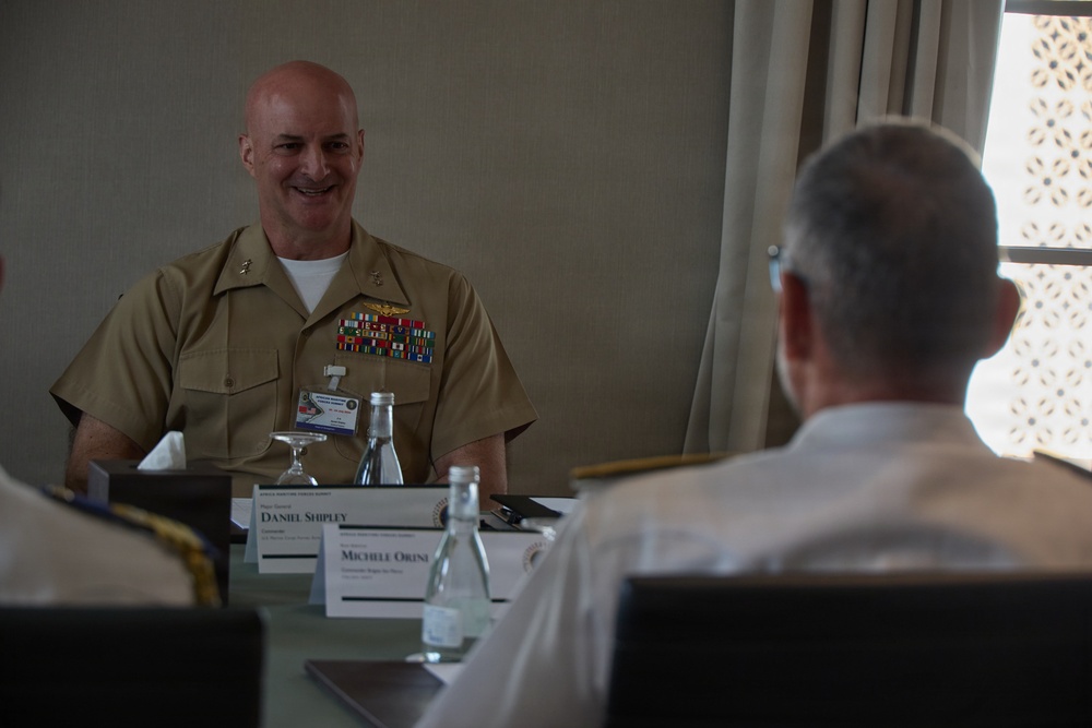 A smiling U.S. Marine Corps officer in tan service uniform with multiple ribbons and decorations is seated at a conference table during what appears to be a formal military summit. Name placards on the table identify attendees including 'Daniel Shipley' and 'Michele Orini', with the event banner reading 'Africa Maritime Forces Summit'. Another uniformed officer is visible from behind in the foreground.