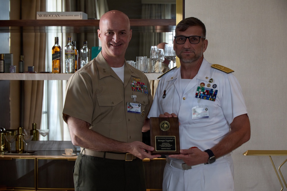 Two military officers are posing together while jointly holding a wooden plaque award. One officer is wearing a tan/khaki U.S. Marine Corps uniform, while the other is wearing a white naval dress uniform with extensive medal ribbons. The plaque appears to read 'Stronger Together' suggesting a formal recognition or commemorative ceremony.