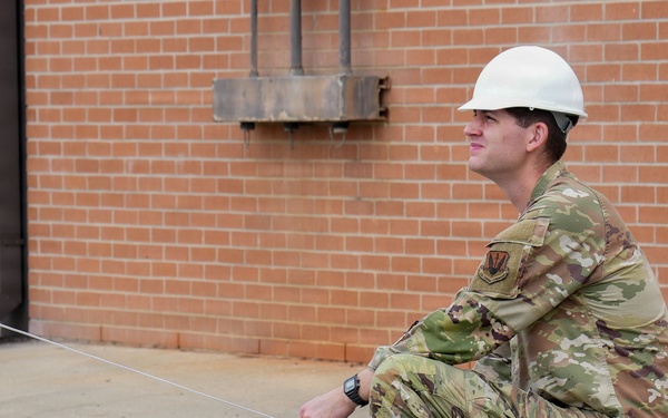 A military service member wearing OCP (Operational Camouflage Pattern) uniform and a white hard hat is crouching on a concrete surface near a brick building. He is holding a taut string or line, likely used for surveying, measuring, or construction layout purposes. The setting appears to be a military installation or construction site.