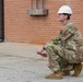 A military service member wearing OCP (Operational Camouflage Pattern) uniform and a white hard hat is crouching on a concrete surface near a brick building. He is holding a taut string or line, likely used for surveying, measuring, or construction layout purposes. The setting appears to be a military installation or construction site.