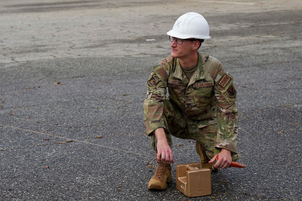 A U.S. Air Force servicemember wearing OCP camouflage uniform and a white hard hat is crouching on a paved surface. The individual, whose name tape reads 'LECHWA' (partial), is handling a small tan box and an orange marker while a rope or cord extends to the left. The scene appears to be an outdoor training or work exercise on a tarmac or parking area.