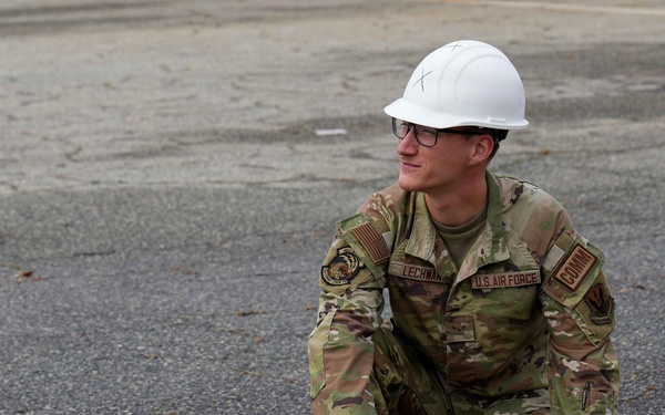 A U.S. Air Force servicemember wearing OCP camouflage uniform and a white hard hat is crouching on a paved surface. The individual, whose name tape reads 'LECHWA' (partial), is handling a small tan box and an orange marker while a rope or cord extends to the left. The scene appears to be an outdoor training or work exercise on a tarmac or parking area.
