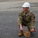 A U.S. Air Force servicemember wearing OCP camouflage uniform and a white hard hat is crouching on a paved surface. The individual, whose name tape reads 'LECHWA' (partial), is handling a small tan box and an orange marker while a rope or cord extends to the left. The scene appears to be an outdoor training or work exercise on a tarmac or parking area.