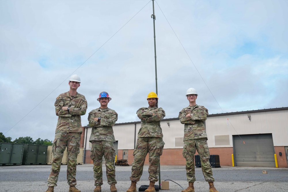 Four military personnel in camouflage uniforms stand in a row with arms crossed in front of a brick building and a tall communications/antenna mast. Three individuals wear hard hats — two white, one yellow, and one patriotic American flag-themed — suggesting a safety-conscious work environment. The setting appears to be a military installation or base facility.