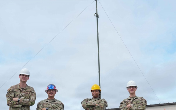 Four military personnel in camouflage uniforms stand in a row with arms crossed in front of a brick building and a tall communications/antenna mast. Three individuals wear hard hats — two white, one yellow, and one patriotic American flag-themed — suggesting a safety-conscious work environment. The setting appears to be a military installation or base facility.