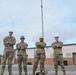 Four military personnel in camouflage uniforms stand in a row with arms crossed in front of a brick building and a tall communications/antenna mast. Three individuals wear hard hats — two white, one yellow, and one patriotic American flag-themed — suggesting a safety-conscious work environment. The setting appears to be a military installation or base facility.