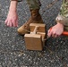 The image shows military personnel in OCP (Operational Camouflage Pattern) uniforms working with what appear to be training demolition blocks or simulated explosive charges on an asphalt surface. One individual is holding a wire or cord connected to the tan-colored rectangular blocks, while another holds an orange tool or marker. The scene appears to be a training exercise involving simulated ordnance placement or EOD (Explosive Ordnance Disposal) procedures.