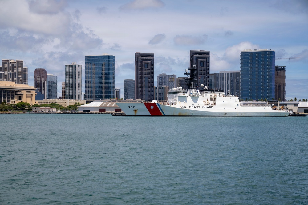 A U.S. Coast Guard cutter bearing hull number 757 is docked at a pier in what appears to be Honolulu, Hawaii, identifiable by the distinctive downtown skyline with high-rise buildings in the background. The vessel is white with the characteristic diagonal red and blue Coast Guard stripe on its bow. The scene is photographed from the water on a partly cloudy day.