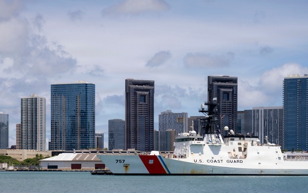 A U.S. Coast Guard cutter bearing hull number 757 is docked at a pier in what appears to be Honolulu, Hawaii, identifiable by the distinctive downtown skyline with high-rise buildings in the background. The vessel is white with the characteristic diagonal red and blue Coast Guard stripe on its bow. The scene is photographed from the water on a partly cloudy day.