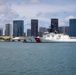 A U.S. Coast Guard cutter bearing hull number 757 is docked at a pier in what appears to be Honolulu, Hawaii, identifiable by the distinctive downtown skyline with high-rise buildings in the background. The vessel is white with the characteristic diagonal red and blue Coast Guard stripe on its bow. The scene is photographed from the water on a partly cloudy day.