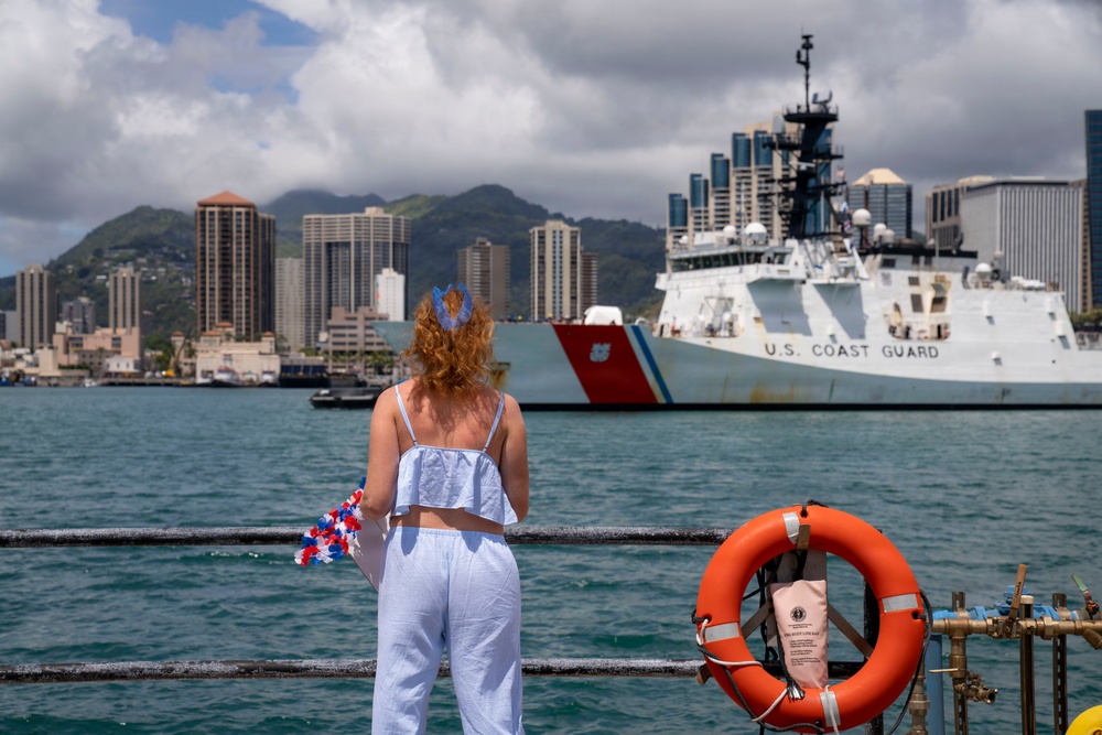 A woman with red/auburn hair stands at a waterfront railing, watching a U.S. Coast Guard cutter vessel approaching or docking in what appears to be Honolulu Harbor, Hawaii. She is holding red, white, and blue decorative flowers or lei, suggesting she is waiting to welcome crew members returning from deployment. The urban skyline of Honolulu and green mountain ridgeline are visible in the background.