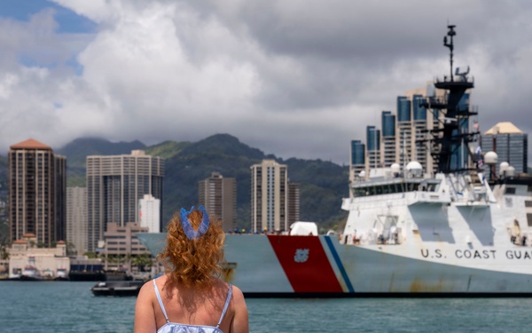 A woman with red/auburn hair stands at a waterfront railing, watching a U.S. Coast Guard cutter vessel approaching or docking in what appears to be Honolulu Harbor, Hawaii. She is holding red, white, and blue decorative flowers or lei, suggesting she is waiting to welcome crew members returning from deployment. The urban skyline of Honolulu and green mountain ridgeline are visible in the background.