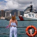A woman with red/auburn hair stands at a waterfront railing, watching a U.S. Coast Guard cutter vessel approaching or docking in what appears to be Honolulu Harbor, Hawaii. She is holding red, white, and blue decorative flowers or lei, suggesting she is waiting to welcome crew members returning from deployment. The urban skyline of Honolulu and green mountain ridgeline are visible in the background.