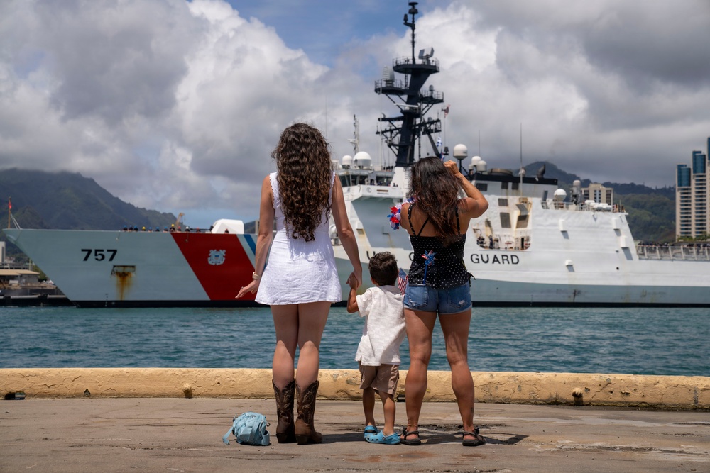 Two women and a young child stand on a pier watching a U.S. Coast Guard cutter (hull number 757) in a harbor. The group appears to be greeting or farewell-waving to the vessel, with one woman waving a patriotic decoration and the child holding a small American flag. The scene is set in what appears to be a tropical port, likely Hawaii, based on the mountainous backdrop and urban skyline.