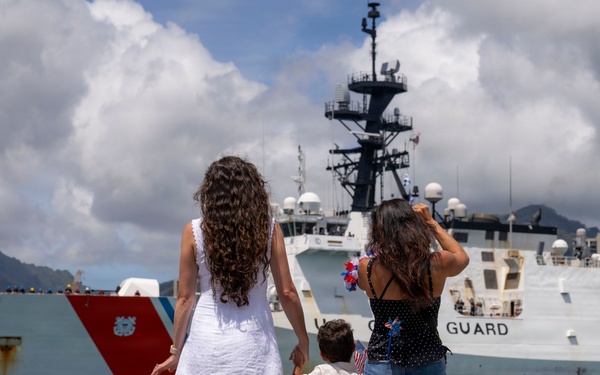 Two women and a young child stand on a pier watching a U.S. Coast Guard cutter (hull number 757) in a harbor. The group appears to be greeting or farewell-waving to the vessel, with one woman waving a patriotic decoration and the child holding a small American flag. The scene is set in what appears to be a tropical port, likely Hawaii, based on the mountainous backdrop and urban skyline.