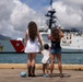 Two women and a young child stand on a pier watching a U.S. Coast Guard cutter (hull number 757) in a harbor. The group appears to be greeting or farewell-waving to the vessel, with one woman waving a patriotic decoration and the child holding a small American flag. The scene is set in what appears to be a tropical port, likely Hawaii, based on the mountainous backdrop and urban skyline.
