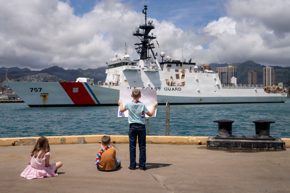 Three children stand and sit on a dock watching a U.S. Coast Guard cutter (hull number 757) arriving or departing at a harbor. The standing child holds up a large sign or photograph, likely greeting a family member aboard the vessel. The setting appears to be a tropical port, consistent with Honolulu, Hawaii, based on the visible skyline and mountainous terrain in the background.