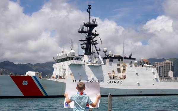 Three children stand and sit on a dock watching a U.S. Coast Guard cutter (hull number 757) arriving or departing at a harbor. The standing child holds up a large sign or photograph, likely greeting a family member aboard the vessel. The setting appears to be a tropical port, consistent with Honolulu, Hawaii, based on the visible skyline and mountainous terrain in the background.