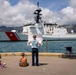 Three children stand and sit on a dock watching a U.S. Coast Guard cutter (hull number 757) arriving or departing at a harbor. The standing child holds up a large sign or photograph, likely greeting a family member aboard the vessel. The setting appears to be a tropical port, consistent with Honolulu, Hawaii, based on the visible skyline and mountainous terrain in the background.