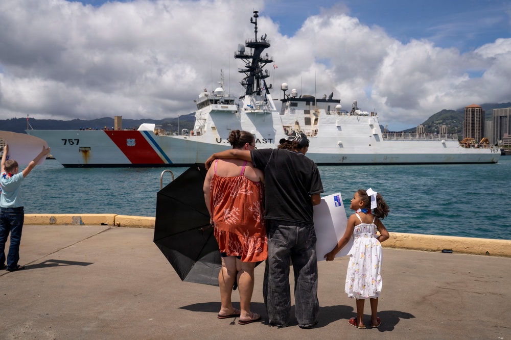 A family stands on a dock watching a U.S. Coast Guard cutter (hull number 757) in a harbor, likely during a departure or homecoming ceremony. Two adults embrace while a young girl in a white dress stands beside them holding a sign. The setting appears to be a tropical port, consistent with Hawaii based on the mountainous backdrop and urban skyline.