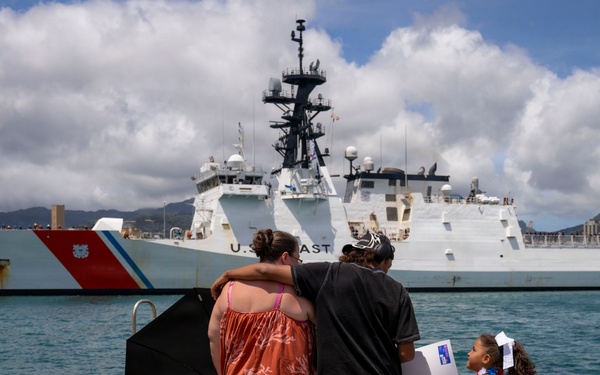 A family stands on a dock watching a U.S. Coast Guard cutter (hull number 757) in a harbor, likely during a departure or homecoming ceremony. Two adults embrace while a young girl in a white dress stands beside them holding a sign. The setting appears to be a tropical port, consistent with Hawaii based on the mountainous backdrop and urban skyline.