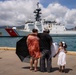 A family stands on a dock watching a U.S. Coast Guard cutter (hull number 757) in a harbor, likely during a departure or homecoming ceremony. Two adults embrace while a young girl in a white dress stands beside them holding a sign. The setting appears to be a tropical port, consistent with Hawaii based on the mountainous backdrop and urban skyline.