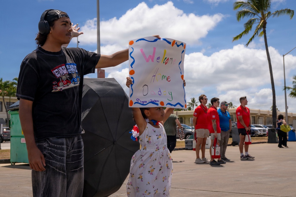 A young girl holds up a handmade 'Welcome Home Daddy' sign decorated with colorful paint and orange dot borders, while a teenage boy stands beside her shielding his eyes from the sun. The scene appears to be a military homecoming event, likely at a base in Hawaii given the palm trees and tropical setting. A group of people in red shirts can be seen in the background, consistent with a welcome home ceremony.