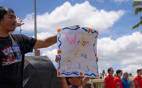 A young girl holds up a handmade 'Welcome Home Daddy' sign decorated with colorful paint and orange dot borders, while a teenage boy stands beside her shielding his eyes from the sun. The scene appears to be a military homecoming event, likely at a base in Hawaii given the palm trees and tropical setting. A group of people in red shirts can be seen in the background, consistent with a welcome home ceremony.