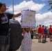 A young girl holds up a handmade 'Welcome Home Daddy' sign decorated with colorful paint and orange dot borders, while a teenage boy stands beside her shielding his eyes from the sun. The scene appears to be a military homecoming event, likely at a base in Hawaii given the palm trees and tropical setting. A group of people in red shirts can be seen in the background, consistent with a welcome home ceremony.