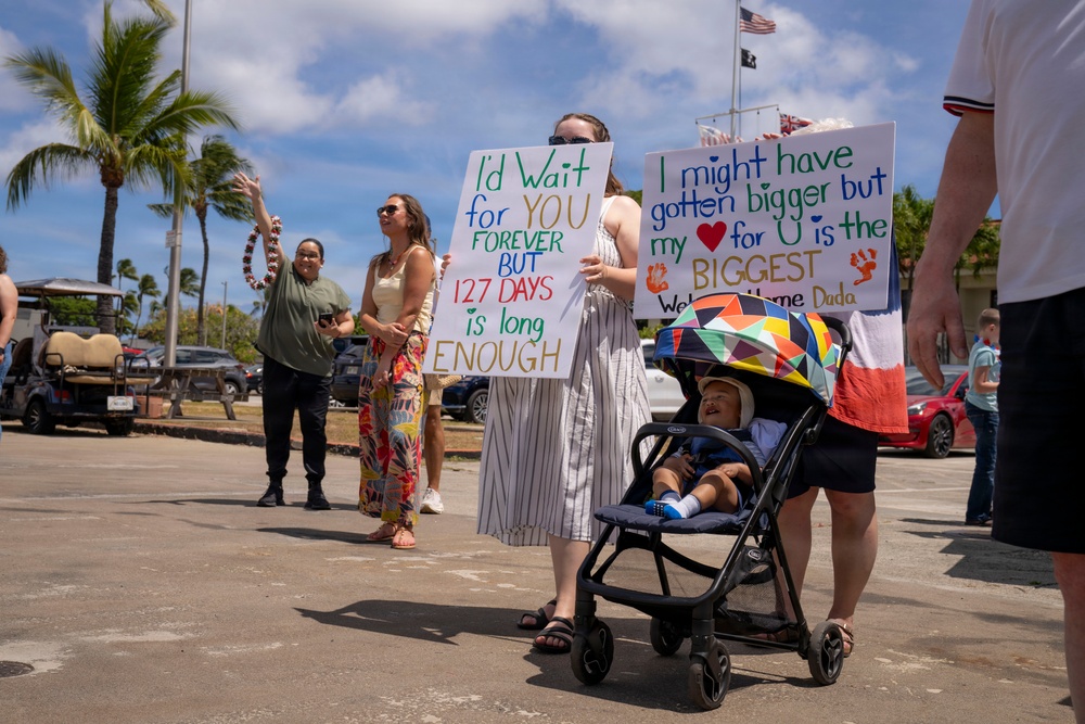A military homecoming or deployment return event, likely in Hawaii based on the palm trees and tropical setting. Family members hold handmade welcome signs, including one reading 'I'd Wait for YOU FOREVER BUT 127 DAYS is long ENOUGH' and another reading 'I might have gotten bigger but my heart for U is the BIGGEST - Welcome Home Dada.' A baby in a stroller and several adults are gathered in what appears to be a parking area.