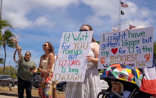 A military homecoming or deployment return event, likely in Hawaii based on the palm trees and tropical setting. Family members hold handmade welcome signs, including one reading 'I'd Wait for YOU FOREVER BUT 127 DAYS is long ENOUGH' and another reading 'I might have gotten bigger but my heart for U is the BIGGEST - Welcome Home Dada.' A baby in a stroller and several adults are gathered in what appears to be a parking area.