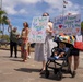 A military homecoming or deployment return event, likely in Hawaii based on the palm trees and tropical setting. Family members hold handmade welcome signs, including one reading 'I'd Wait for YOU FOREVER BUT 127 DAYS is long ENOUGH' and another reading 'I might have gotten bigger but my heart for U is the BIGGEST - Welcome Home Dada.' A baby in a stroller and several adults are gathered in what appears to be a parking area.