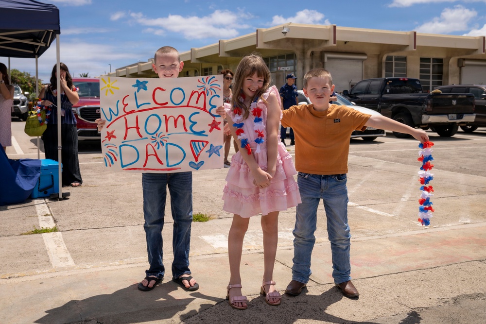 Three children stand together in a parking lot holding a handmade 'Welcome Home Dad' sign and red, white, and blue lei garlands, suggesting a military homecoming event. The children are smiling and appear excited, dressed in casual and festive attire. A uniformed Coast Guard personnel is visible in the background near a building.